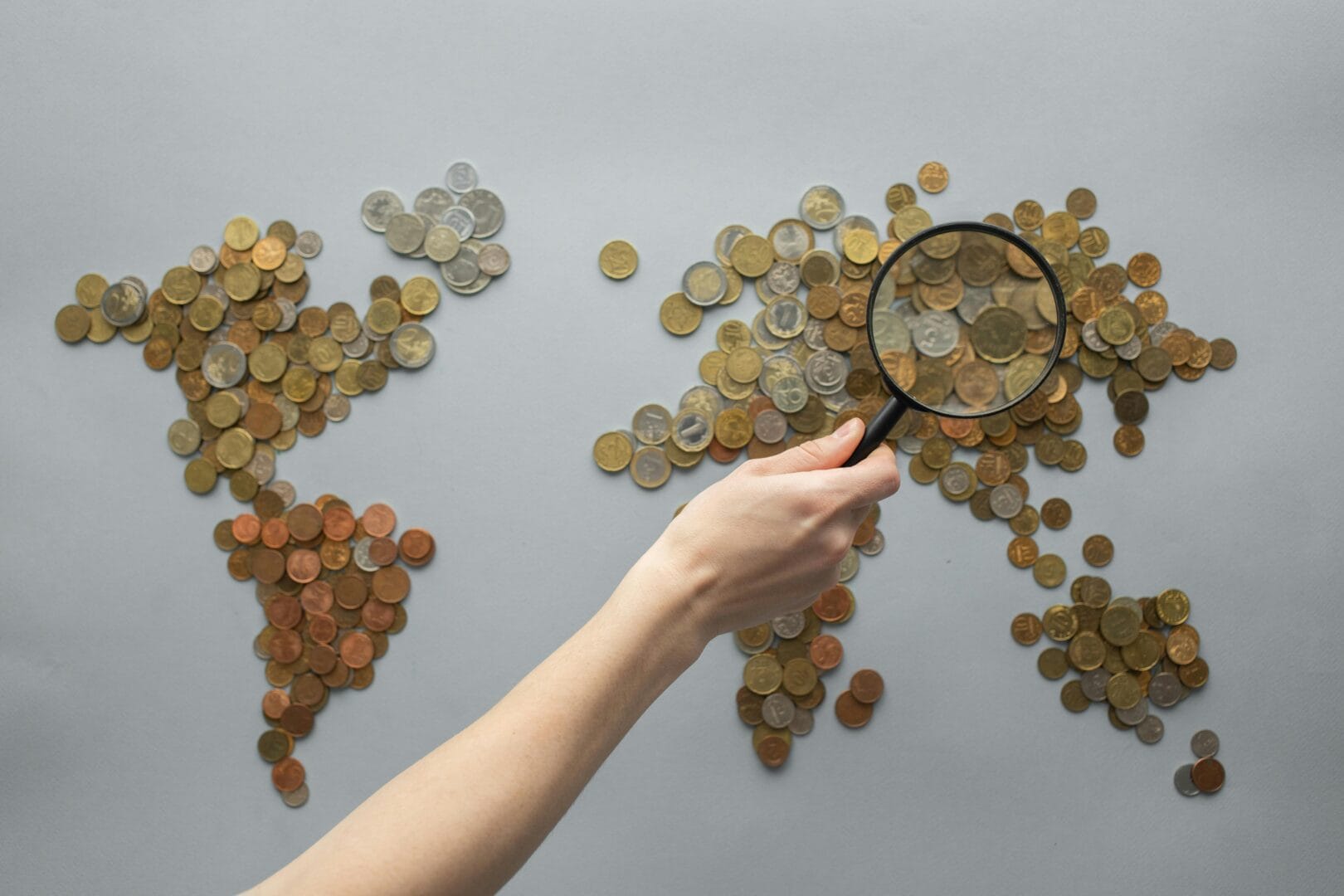 Hand holding a magnifying glass over a map of the world made of coins.