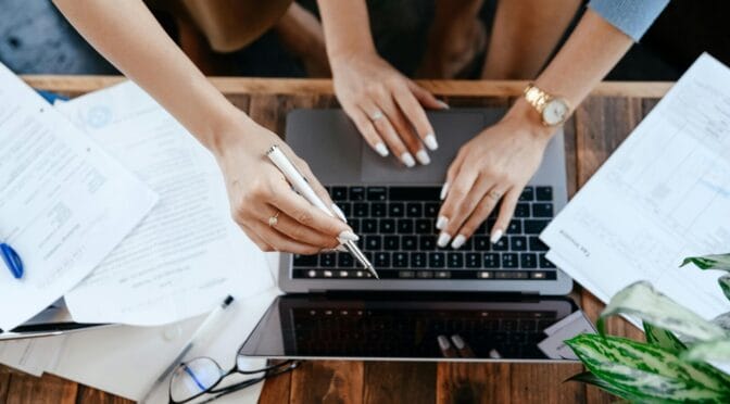 Two people collaborating over a laptop with papers and a plant on a wooden table.