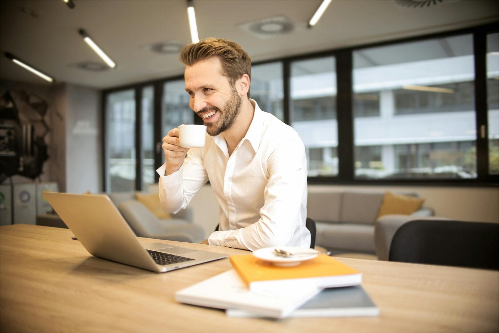 Man in a white shirt sitting at a desk with a laptop, smiling and holding a cup of coffee.